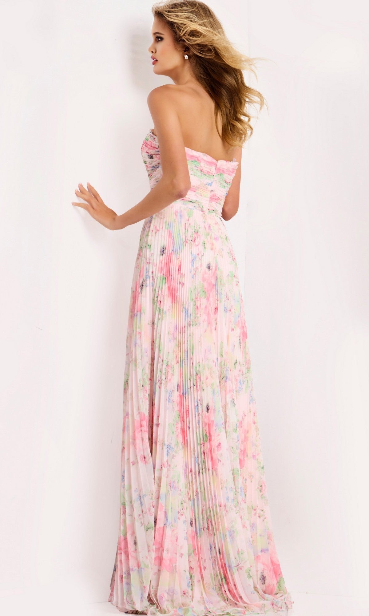 A woman stands with her back to the camera against a plain white background, showcasing the Jovani 44518 long prom dress with a strapless pastel floral design and floor-length pleated skirt.